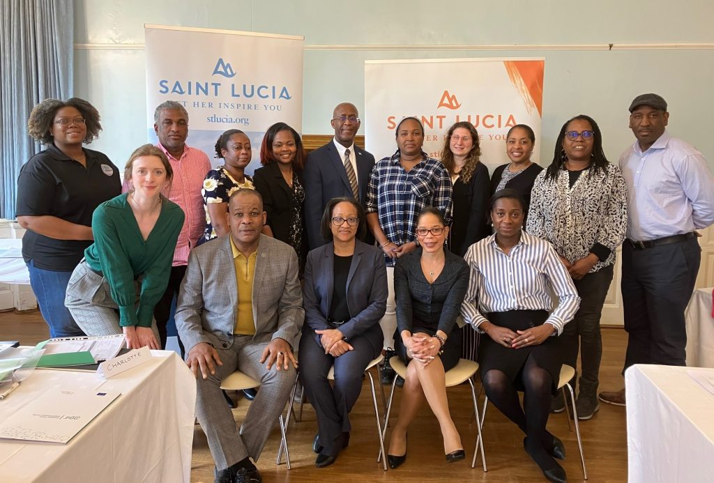 A group of fifteen women and men smile at the camera. They are standing in front of two promotional banners for Saint Lucia.