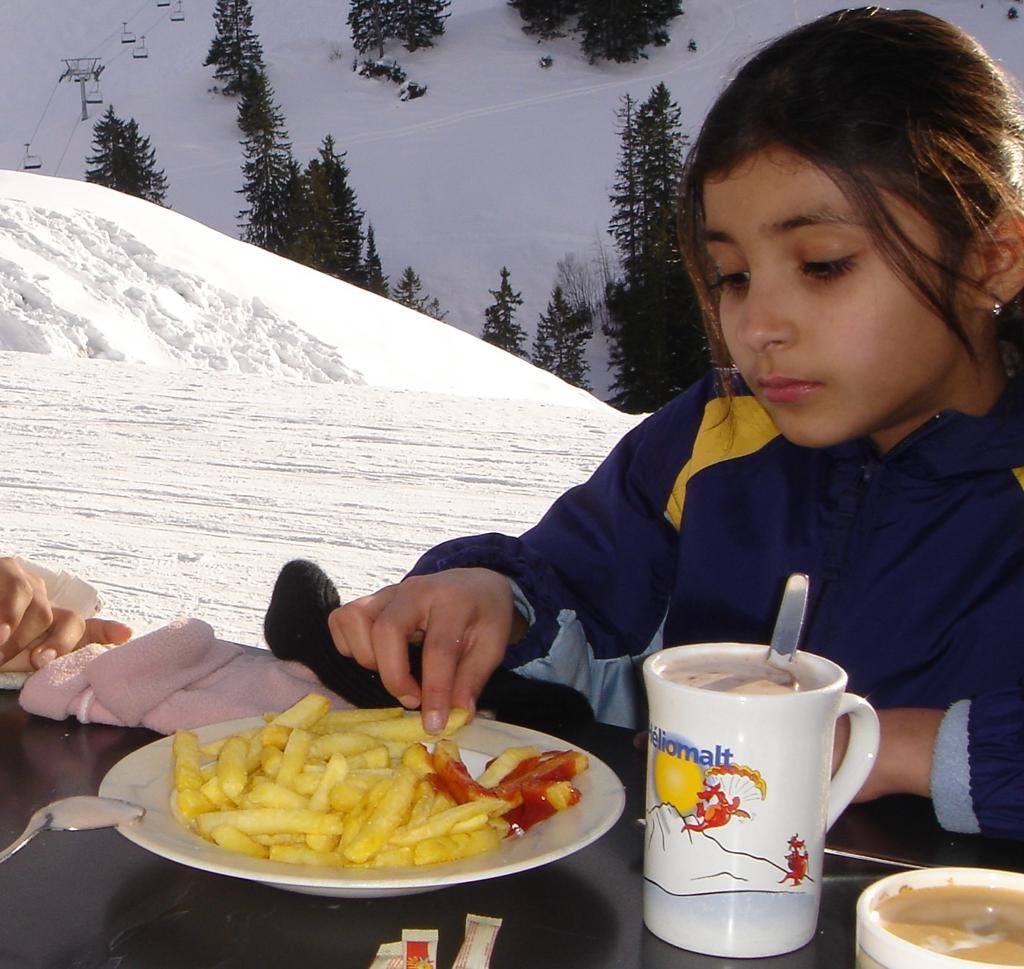 A young girl eating fries with ketchup and drinking hot chocolate in the Alps. 