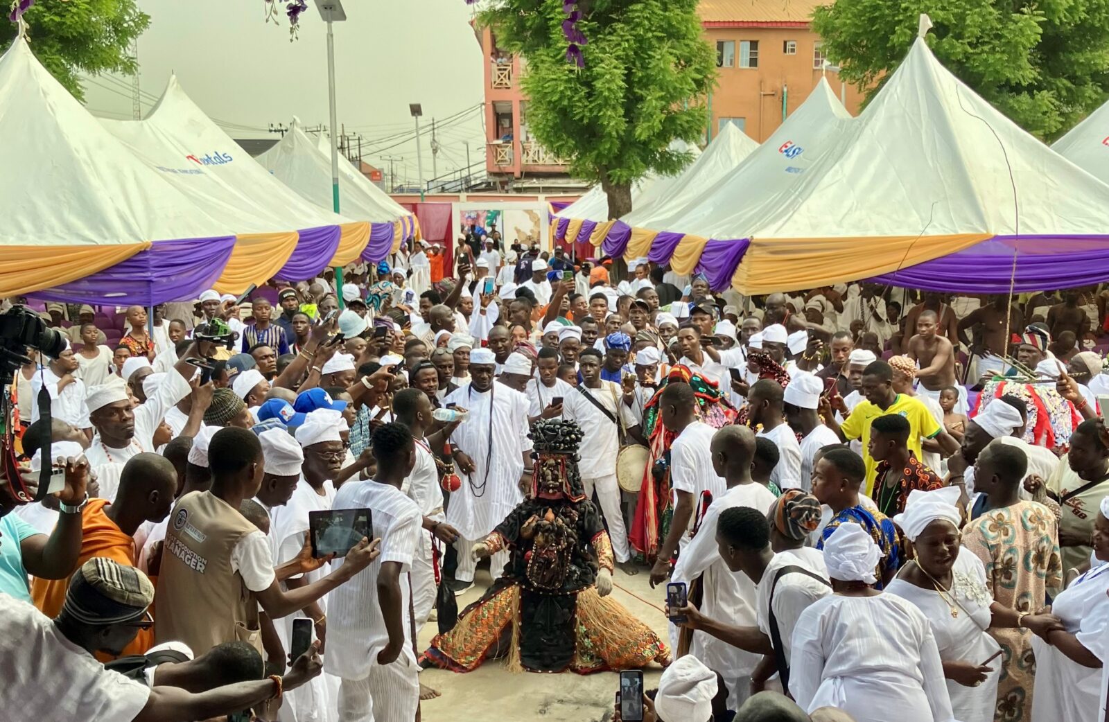 A festival in Ikorodo, Lagos, Nigeria. A figure dressed in traditional costume is on their knees with a large crowd of people surrounding them taking photos.