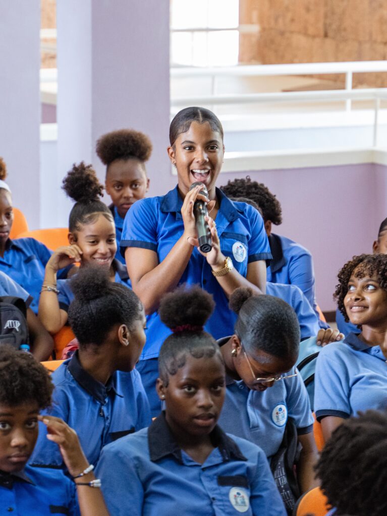 A girl in a uniform stands with a microphone and speaks. Around her there are a several other female students looking at her.