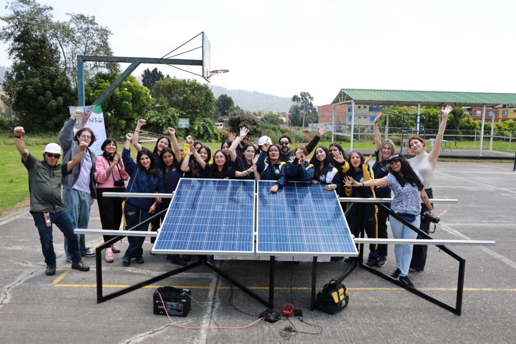 Image of a group of school girls and teachers standing around a solar panel with their hands raised in celebration. 
