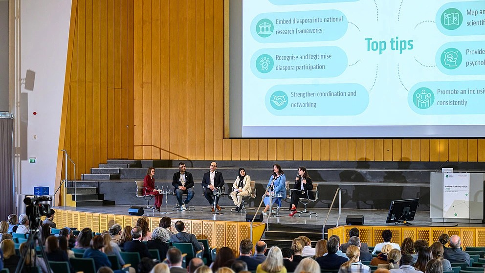 Picture of a panel at an event with 5 people on stage and multiple people in the audience.